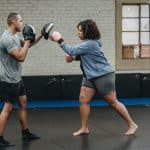 focused black plump female punching boxing paw of ethnic trainer